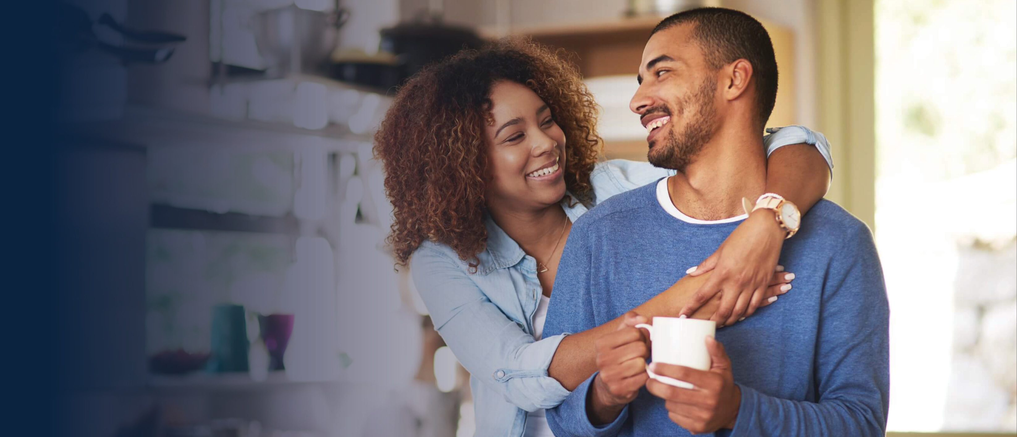 Couple in their kitchen, woman stands behind husband with arms around him.