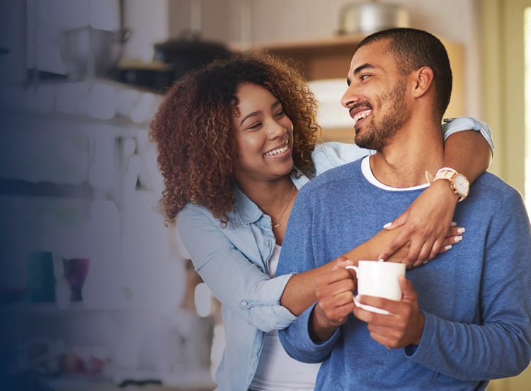 Couple in their kitchen, woman stands behind husband with arms around him.